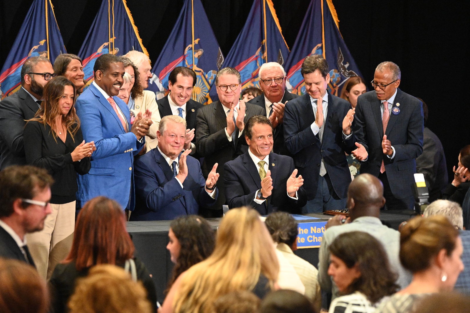 A group of people in suits stand around a table in front of an audience while clapping