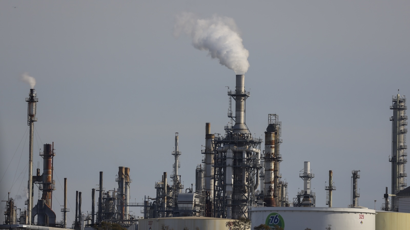 Smoke pours from a refinery smokestack, with gray-blue sky in background.