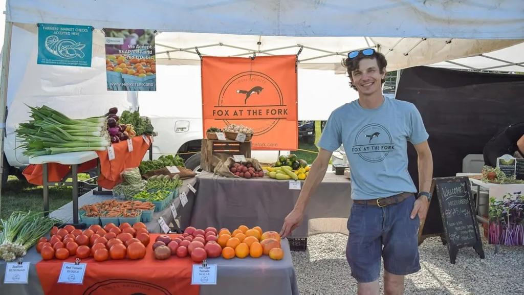 A young white man in denim shorts stands in front of a produce stand at a farmers market.