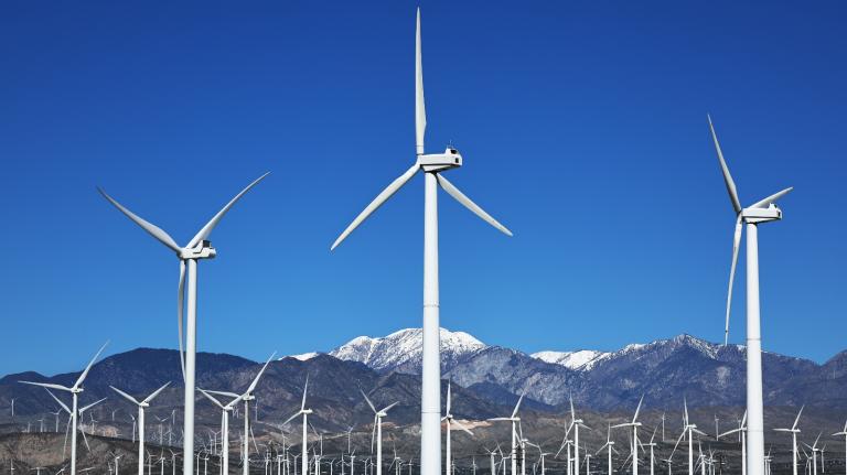 A series of white wind turbines stand under snow capped mountains