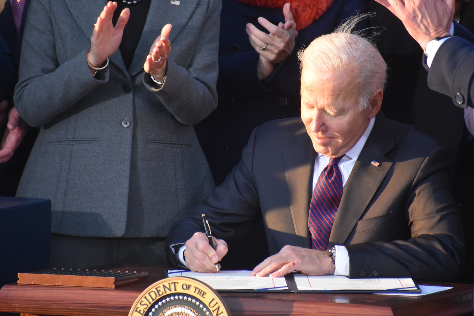 A white-haired man in a suit sits at a desk signing a paper with the part of the U.S. presidential seal visible in front of the desk