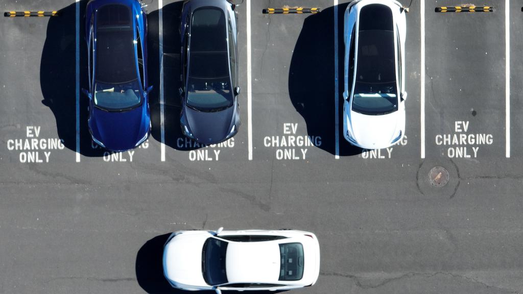 Aeria view of a parking lot, with three cars parked and one in motion. The unclaimed parking spots say 'EV CHARGING ONLY'