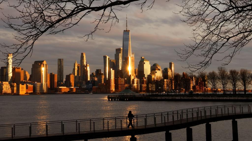 The skyline of lower Manhattan is seen in the distance, the World Trade Center glinting in the sunlight, with water and a bridge on which a person is walking in the foreground