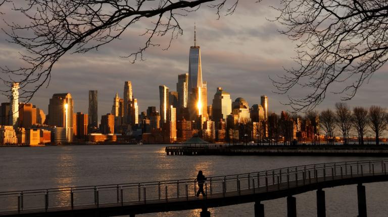 The skyline of lower Manhattan is seen in the distance, the World Trade Center glinting in the sunlight, with water and a bridge on which a person is walking in the foreground