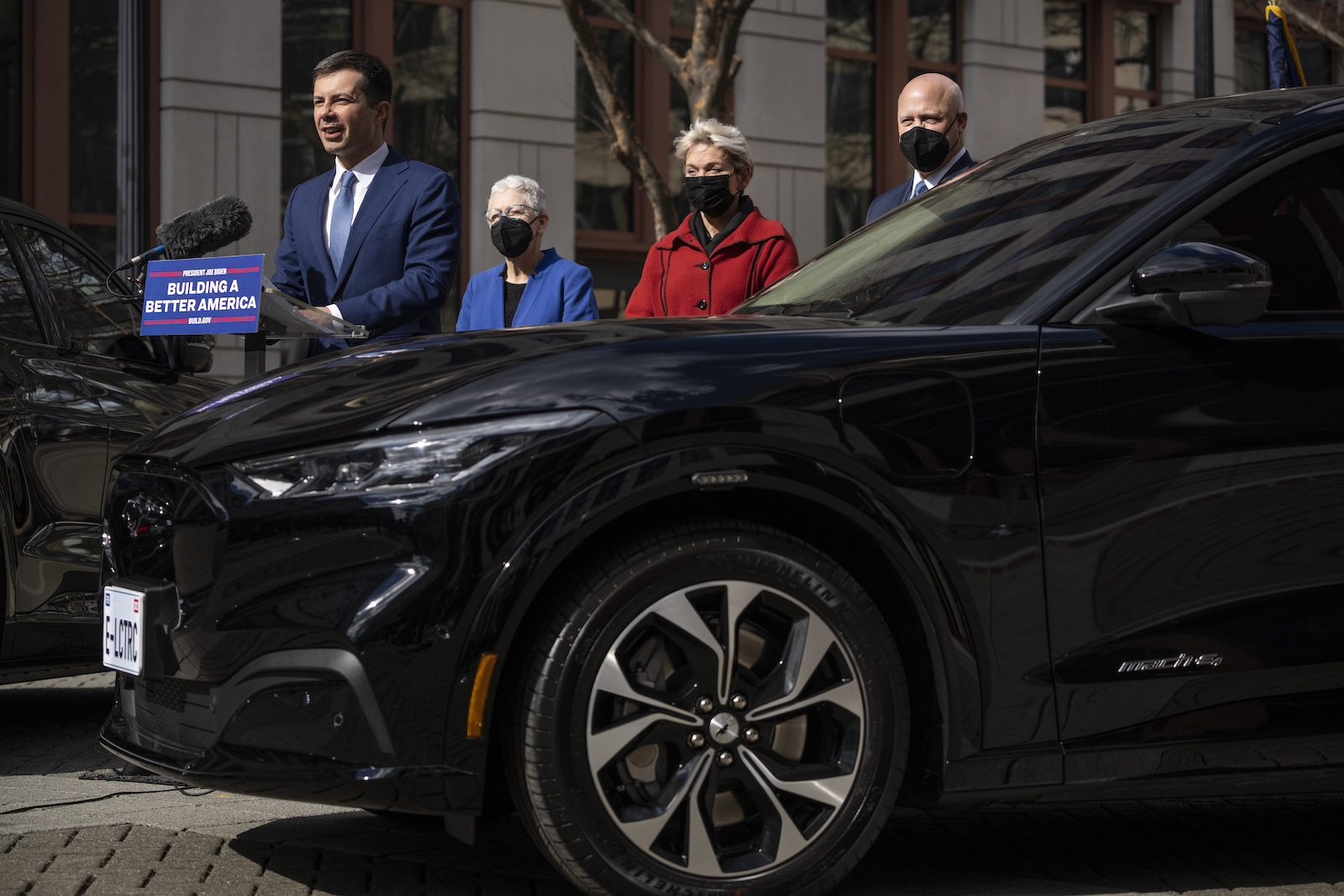 A white man in a suit stands at a podium behind a large black car, with three people wearing masks standing behind him