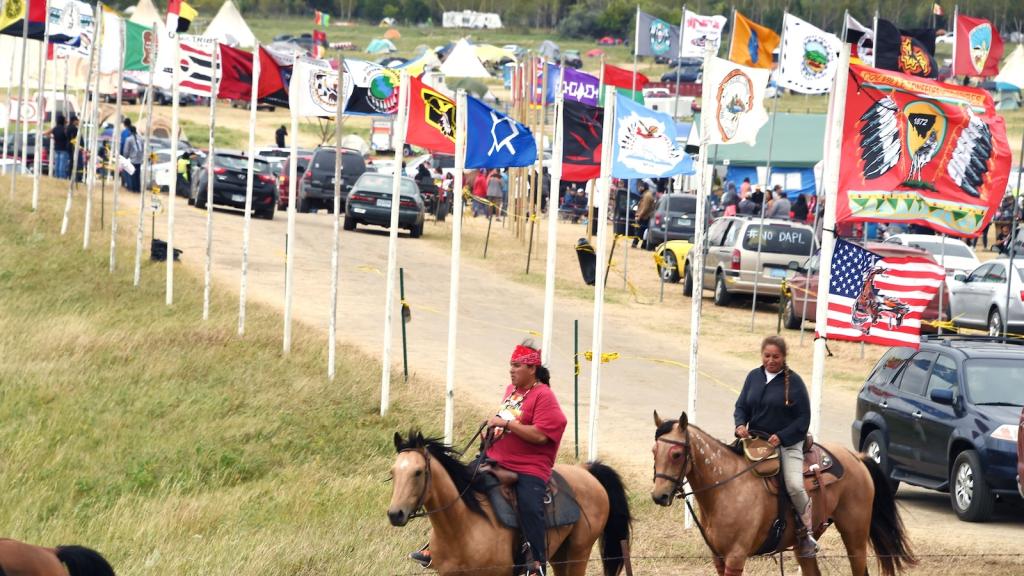 Horseback riders in the foreground, and behind them a dirt road lined by many tribal flags.