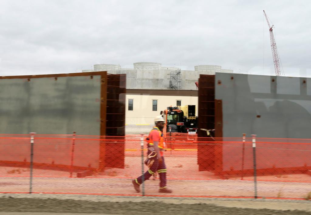 A worker wearing a hard hat and safety vest walks past the cooling towers of a power plant under construction.