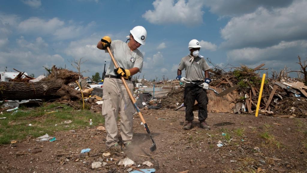 A woman and a man in white hard hats stand in a debris field while one holds a rake