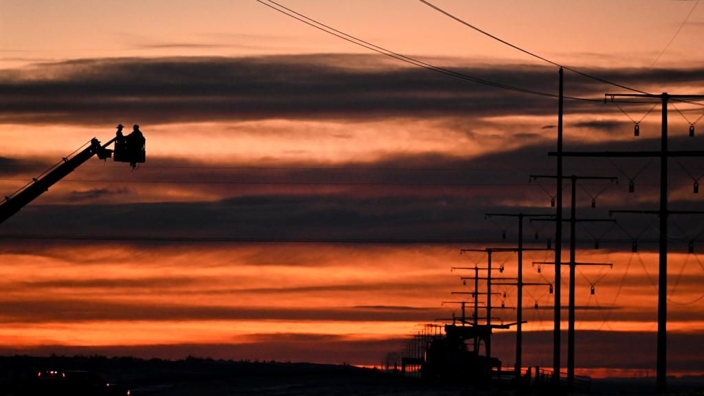 People in the bucket of a boom truck near power poles and lines, with a red and orange sky in the background.