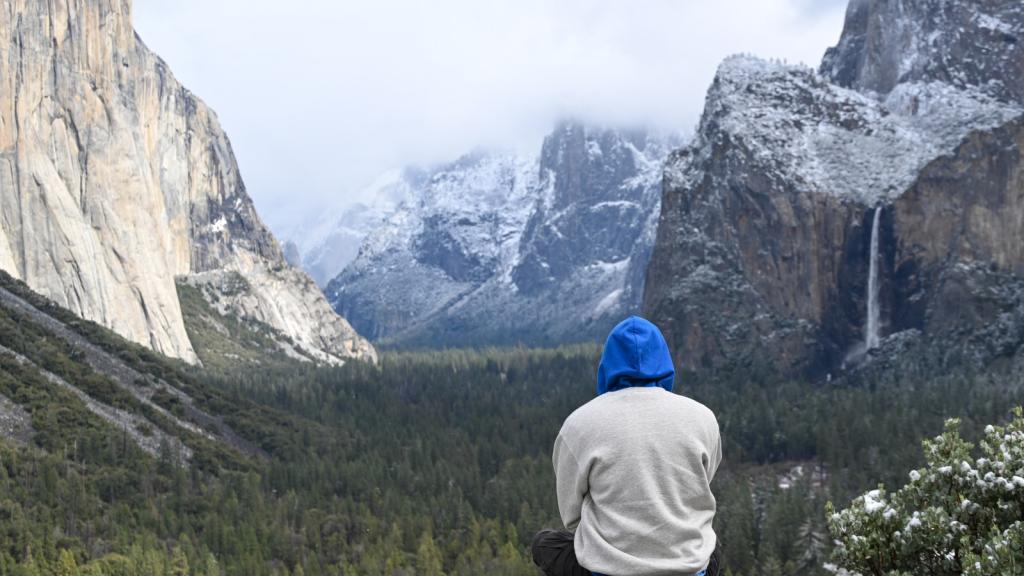 A person in a blue hoodie and sweat shirt looks out on Yosemite valley