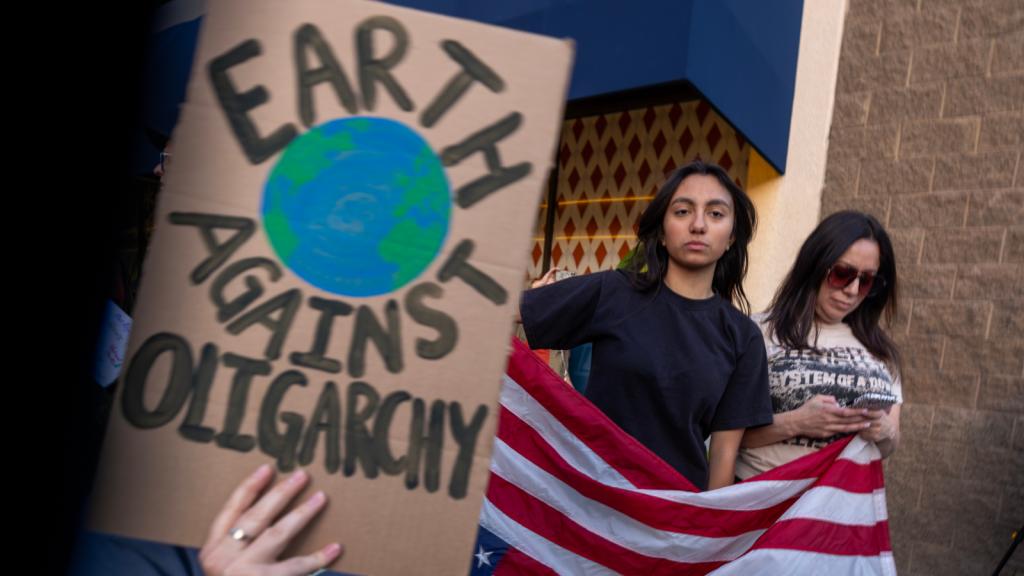 Photo of a protest sign reading Earth Against Oligarchy, with protesters holding flag in the background