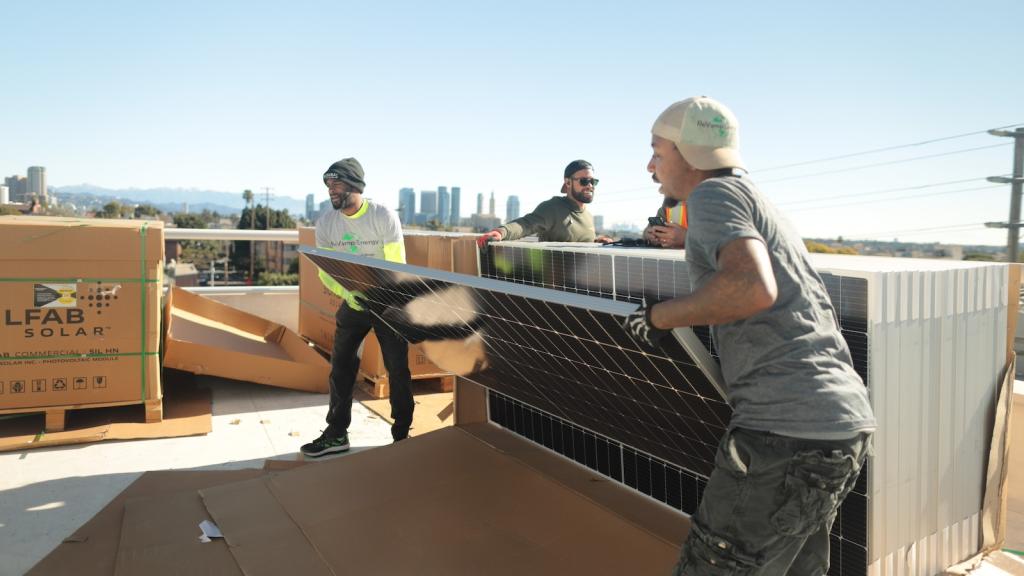 Two men wearing jeans and t-shirts lift a solar panel from the front of a stack of panels on top of a roof, with clear skies behind them