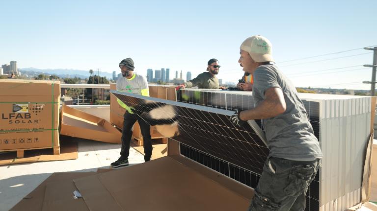Two men wearing jeans and t-shirts lift a solar panel from the front of a stack of panels on top of a roof, with clear skies behind them