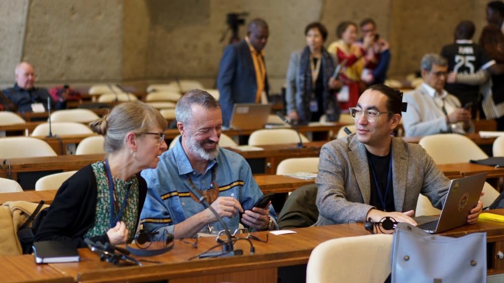 A woman and two men sit at a table and chat with other people talking in the background