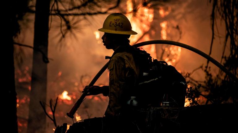 A firefighter in a hard hat holds a hose as a fire blazes in the background