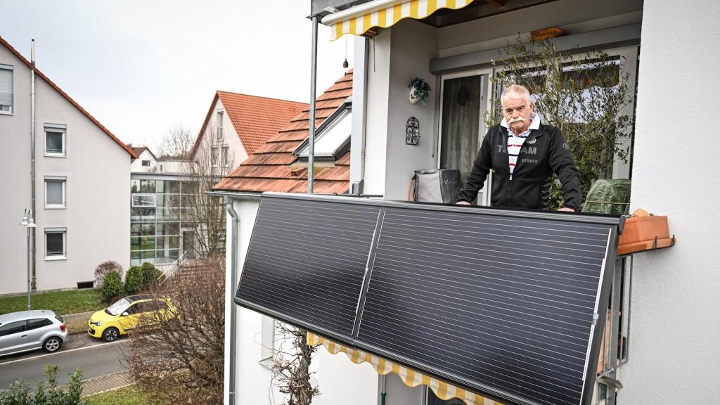 A man stands on his balcony, which features two small solar panels that generate electricity for his apartment.