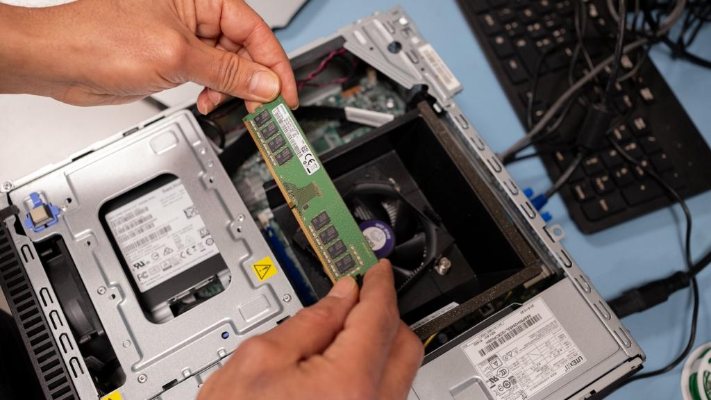 A close-up of someone's hands holding a green strip containing several computer chips above a disassembled computer, next to a black keyboard, on a light blue tabletop