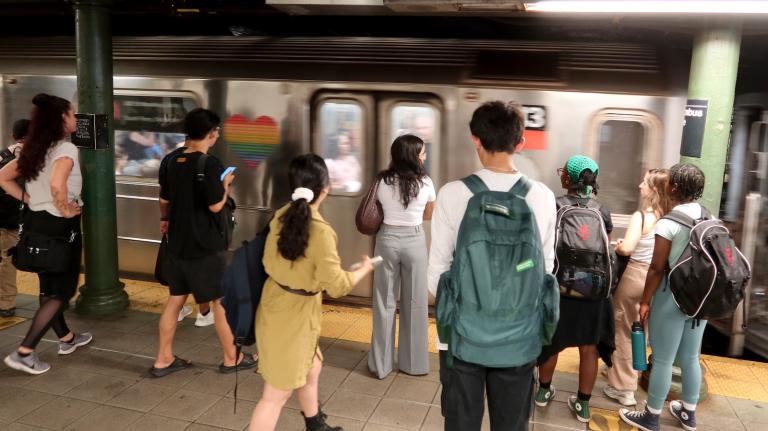 Several people, viewed from behind, stand on a subway platform as a silver subway car appears to pull into the station