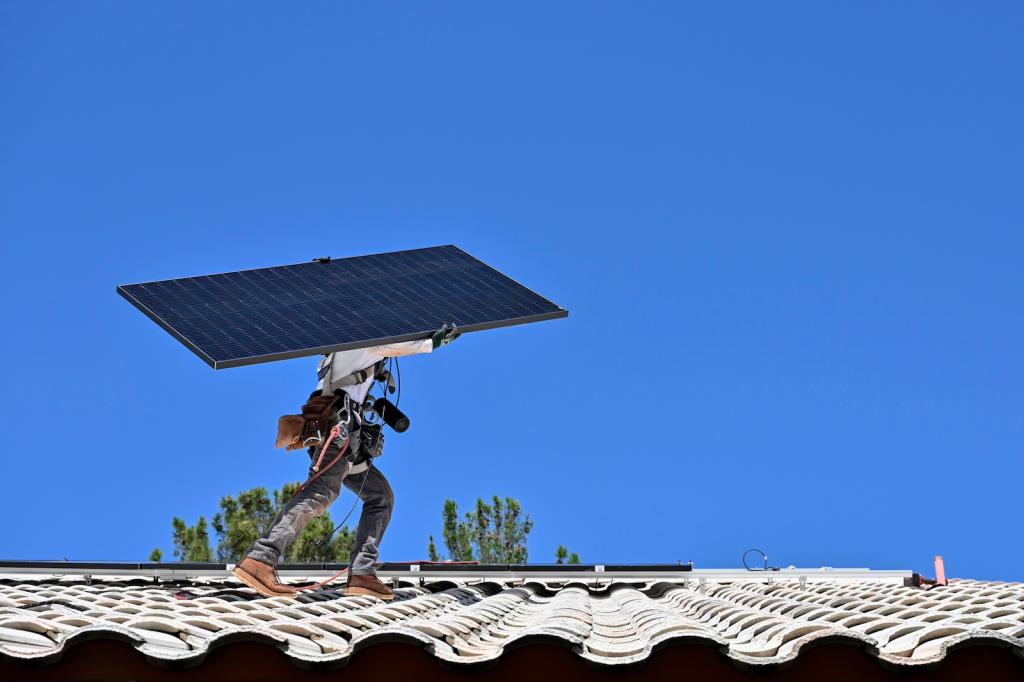 A person wearing jeans and a tool belt carries a solar panel across a roof, with a deep blue sky behind them