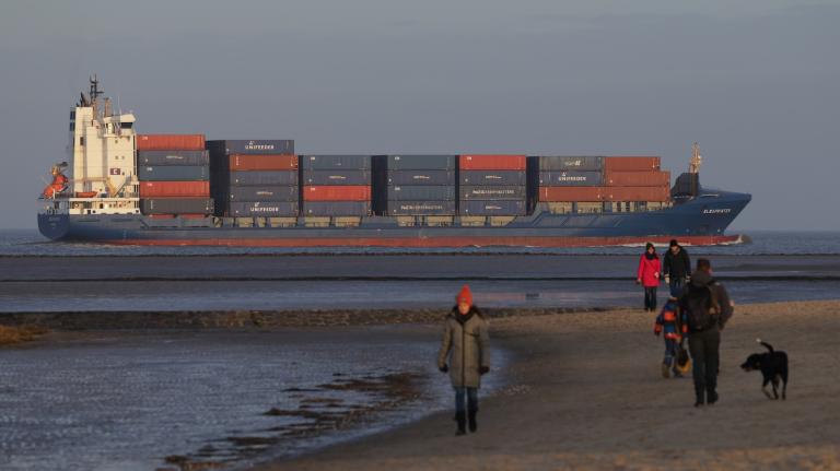 A large freight ship in background, with people walking on the beach in foreground, with hazy air.
