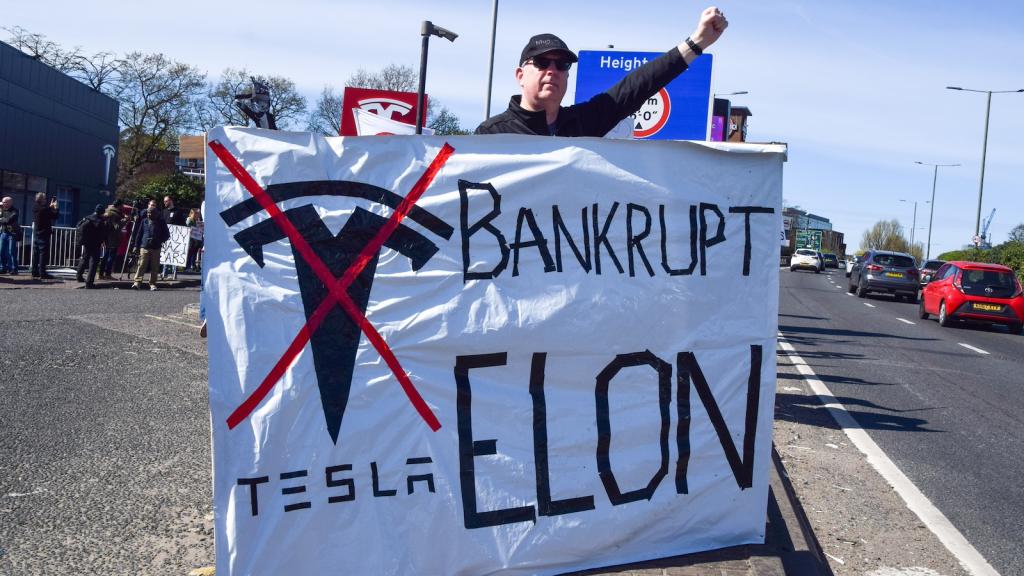 A man in a black shirt and baseball cap stands with his fist raised behind a large banner that reads 'Bankrupt Elon' with an X through the Tesla logo