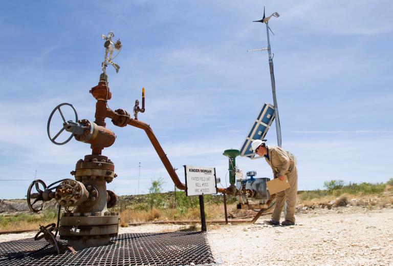 A person stands on a dirt road and inspects a rusty carbon capture well