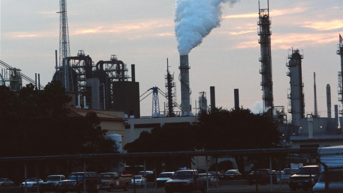 Dark view of a factory with plume emitting from a chimney and cars parked in the parking lot.