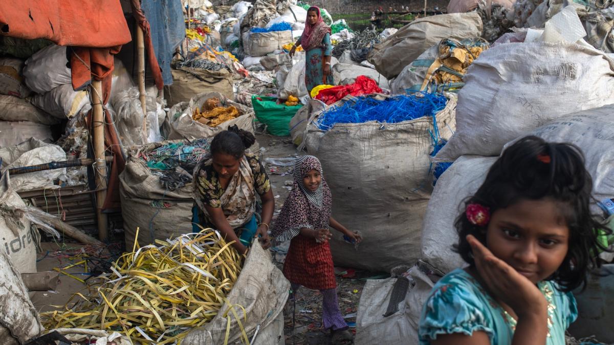 Two children and two adults in a recycling facility in Dhaka, Bangladesh. Bags of shredded plastic fill the room, with some spilling their contents.