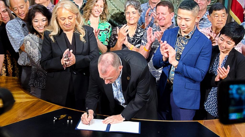A man in a suit signs a piece of paper with people clapping behind him