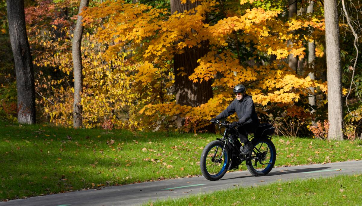 A man rides a bicycle in a park in front of lush fall foliage