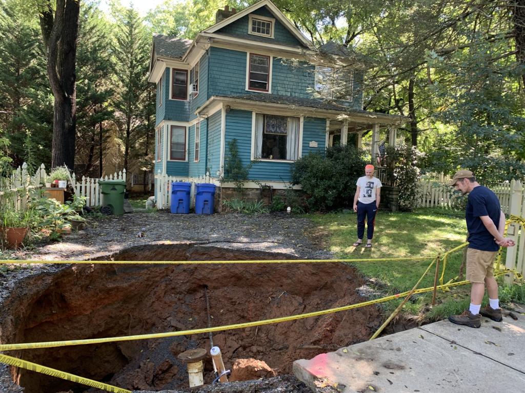 Two people stand near a large sinkhole with caution tape in front of a blue house