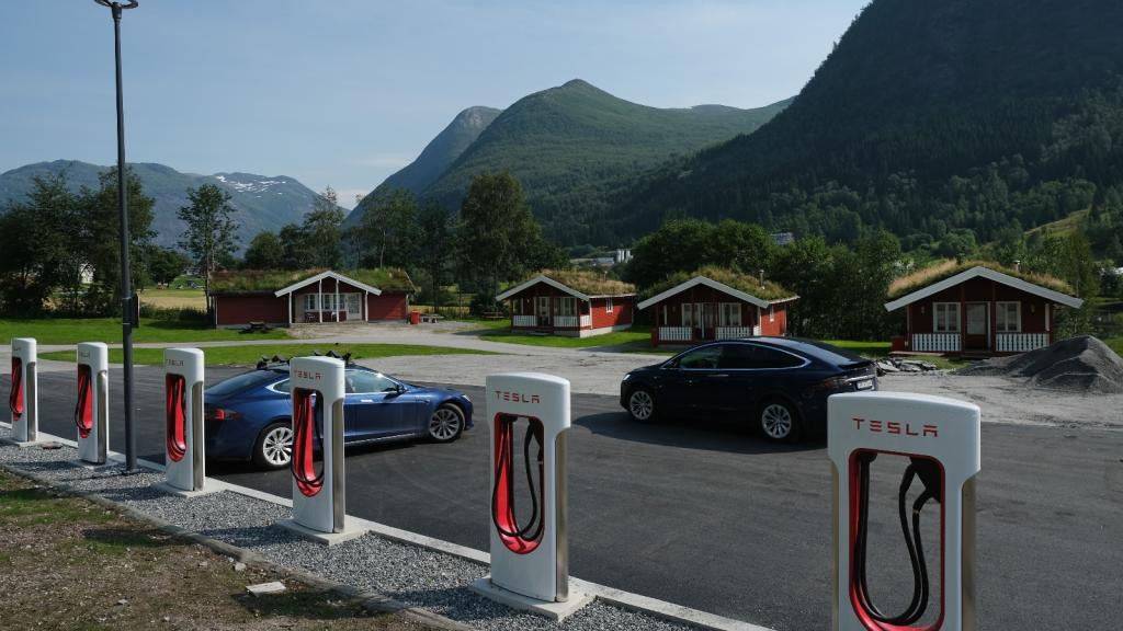 Several electric cars sit next to Tesla chargers in front of idyllic red cabins nestled below green mountains