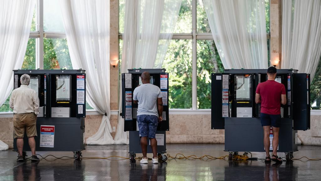 Three men stand in front of three voting machines, their backs to the viewer.