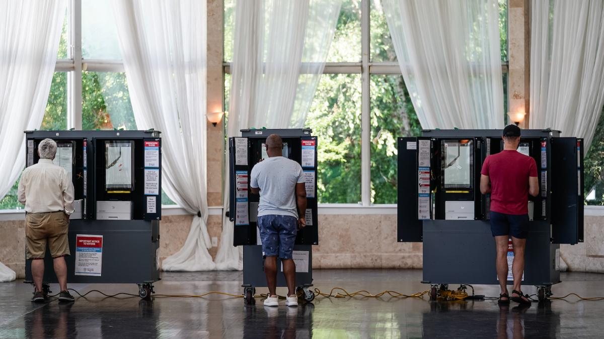 Three men stand in front of three voting machines, their backs to the viewer.