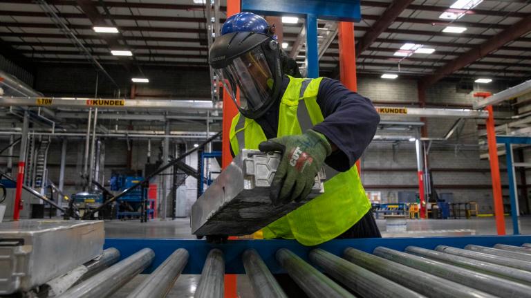 A person wearing a helmet with a face shield, a yellow vest, and gloves places a large, metallic, rectangular prism on an assembly line