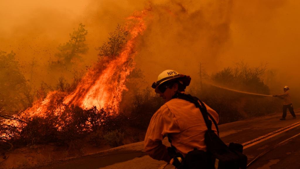 Firefighter stands on road next to a wildfire. The photo is tinged red from fire.