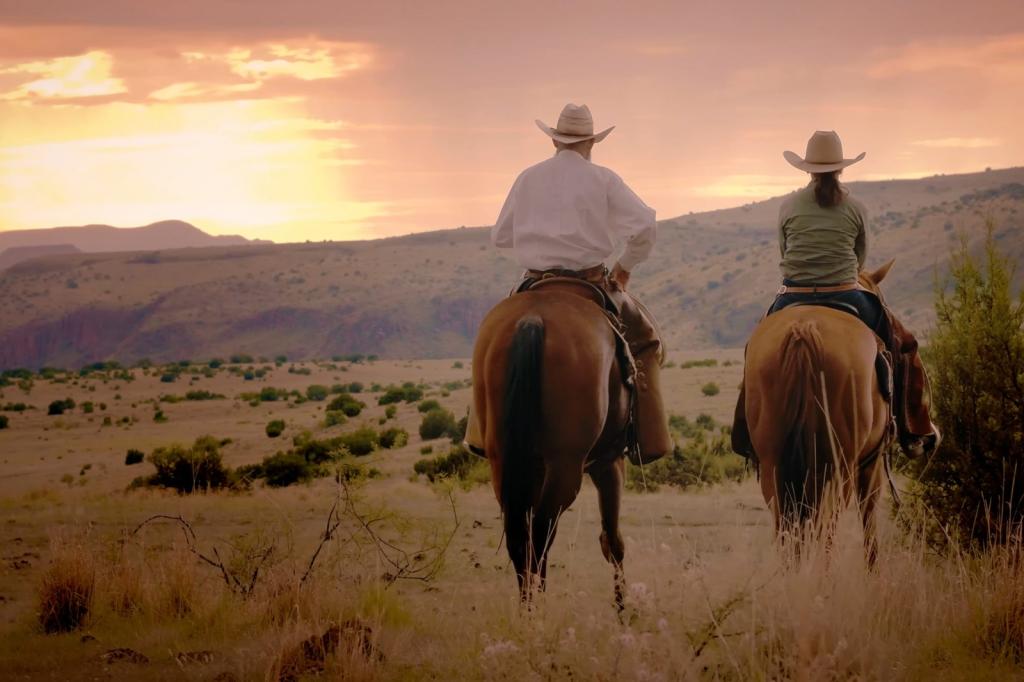 Two people shown from behind wearing cowboy hats and sitting astride horses while looking at the sun set over mountains