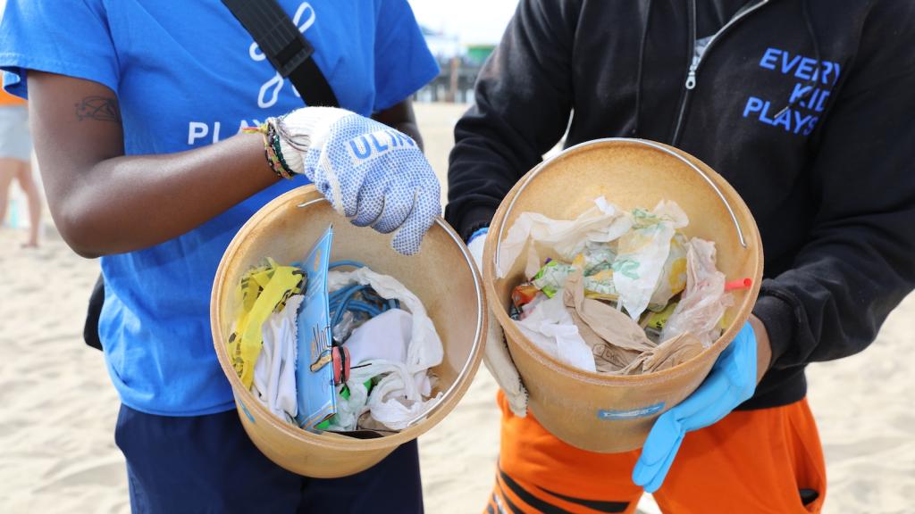 Torsos of two people who are tilting buckets toward the camera. Inside their buckets is trash collected from the beach.