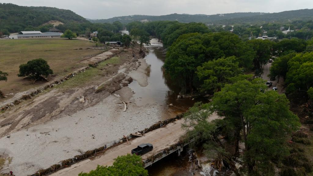 Under gray skies a swollen river flows by trees and a muddy bank