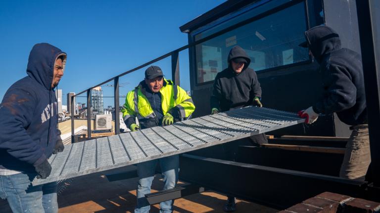 A group of men hold a huge metal grate on top of an apartment building