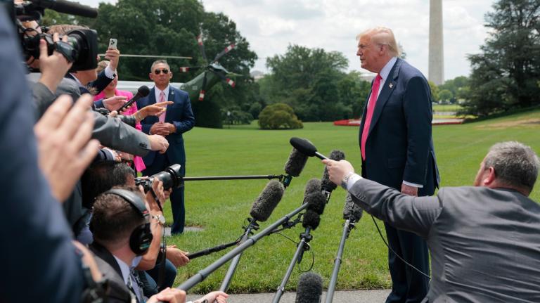 A man in a suit with a red tie and blonde hair stands on a podium with reporters in front of him on a green lawn
