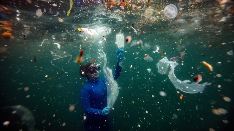 Diver underwater in a blue wetsuit, near the surface, with plastic floating all around them.