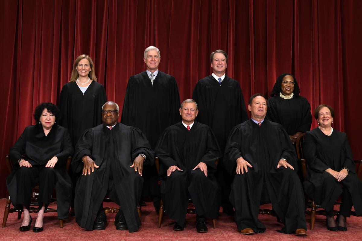 Supreme Court justices pose in two rows, wearing robes, in front of a red curtain.