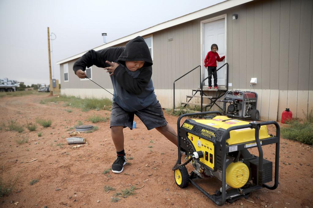 A young boy in a sweatshirt pulls the cord on a yellow generator hooked up to a house nearby while a young girl looks on from the steps
