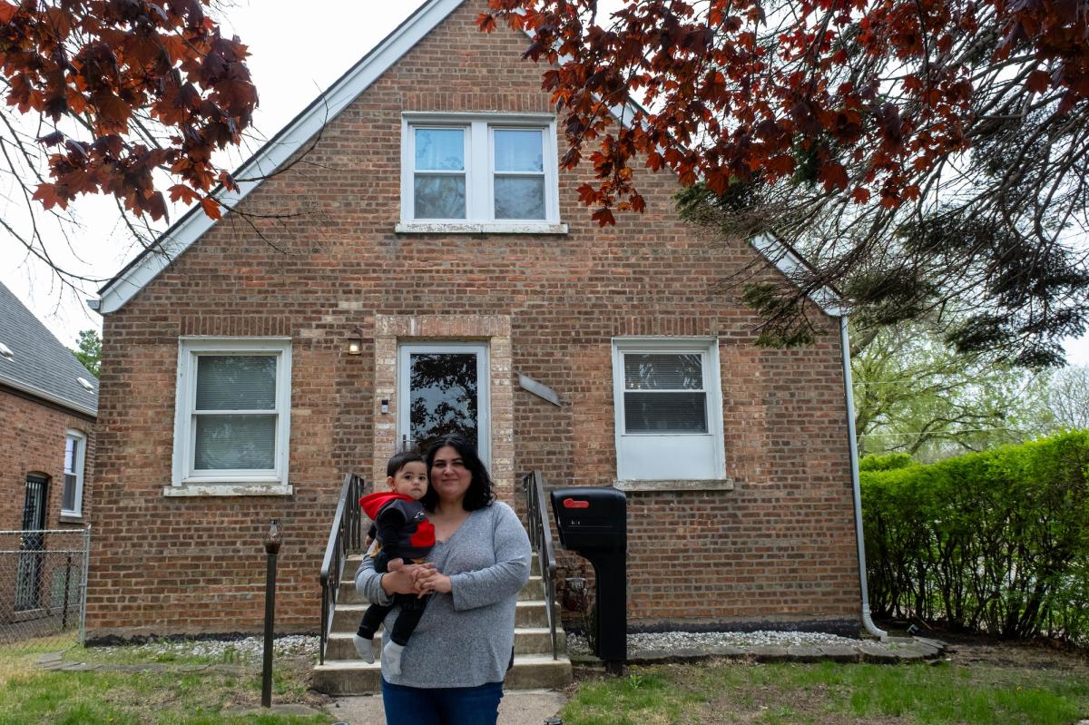 A mother holds a toddler in front of a brick house in a residential area
