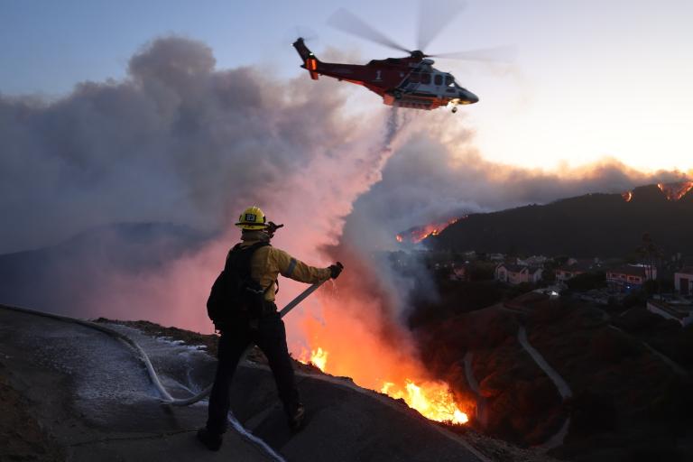 A firefighter stands on a hill overlooking Los Angeles. He's spraying a hose. Below him is a fire and billowing smoke; above him is a helicopter.