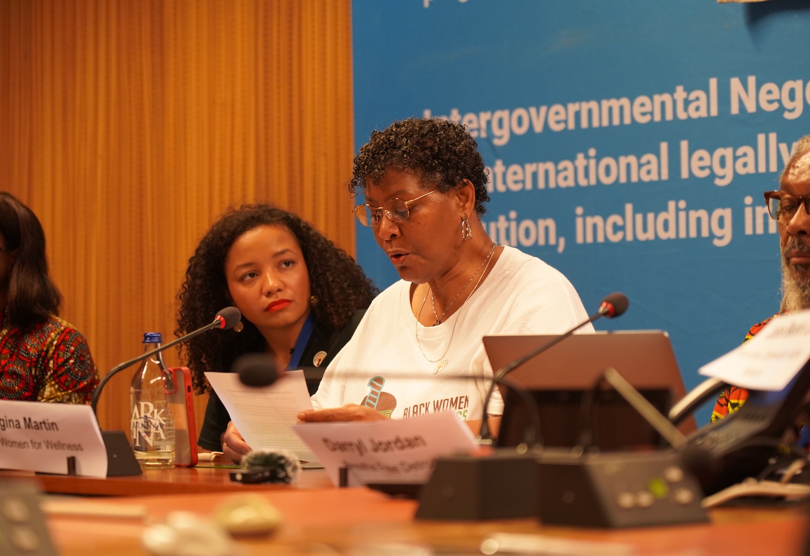 Woman in a white shirt reads from a paper, with microphone in front of her. Another woman sits to her right, and in the background is a blue banner with white text.