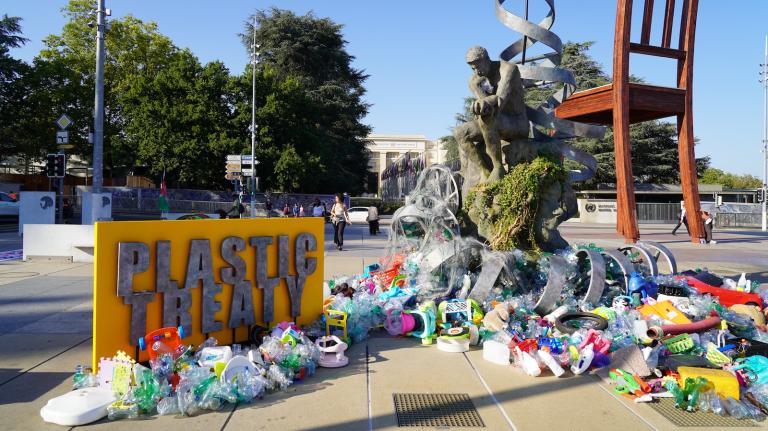 Sculpture of replica of Rodin's The Thinker sitting on a mossy Mother Earth, with plastic pollution at its base. A yellow and gray sign reads 