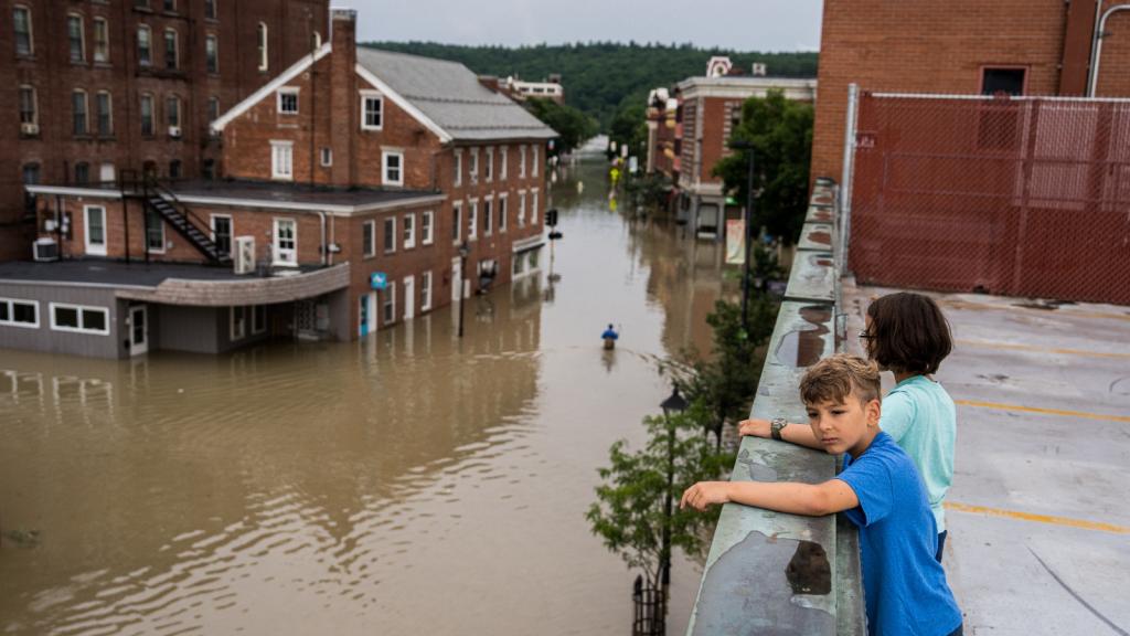 Two boys stand on a roof overlooking a completely flooded street in a pretty red brick town ringed by forests