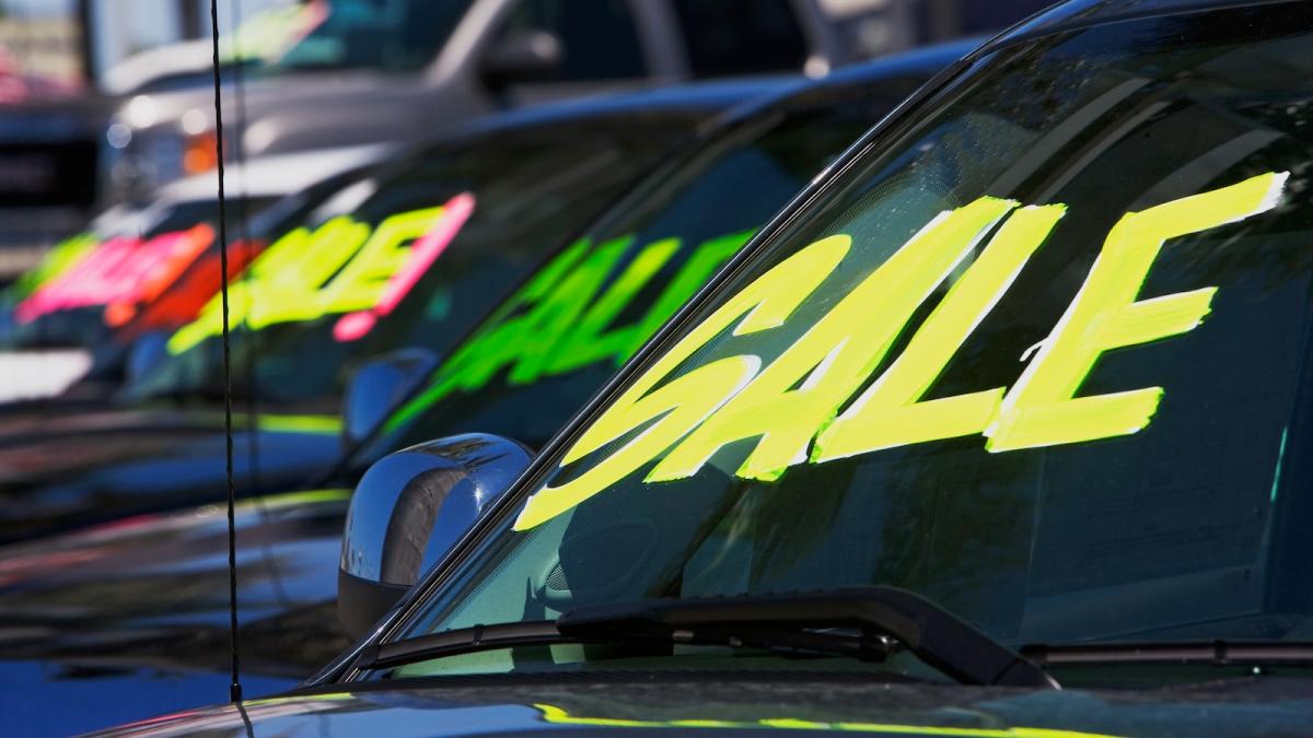 A line of vehicles with "sale" stickers on their windshields is shown at a car dealership.